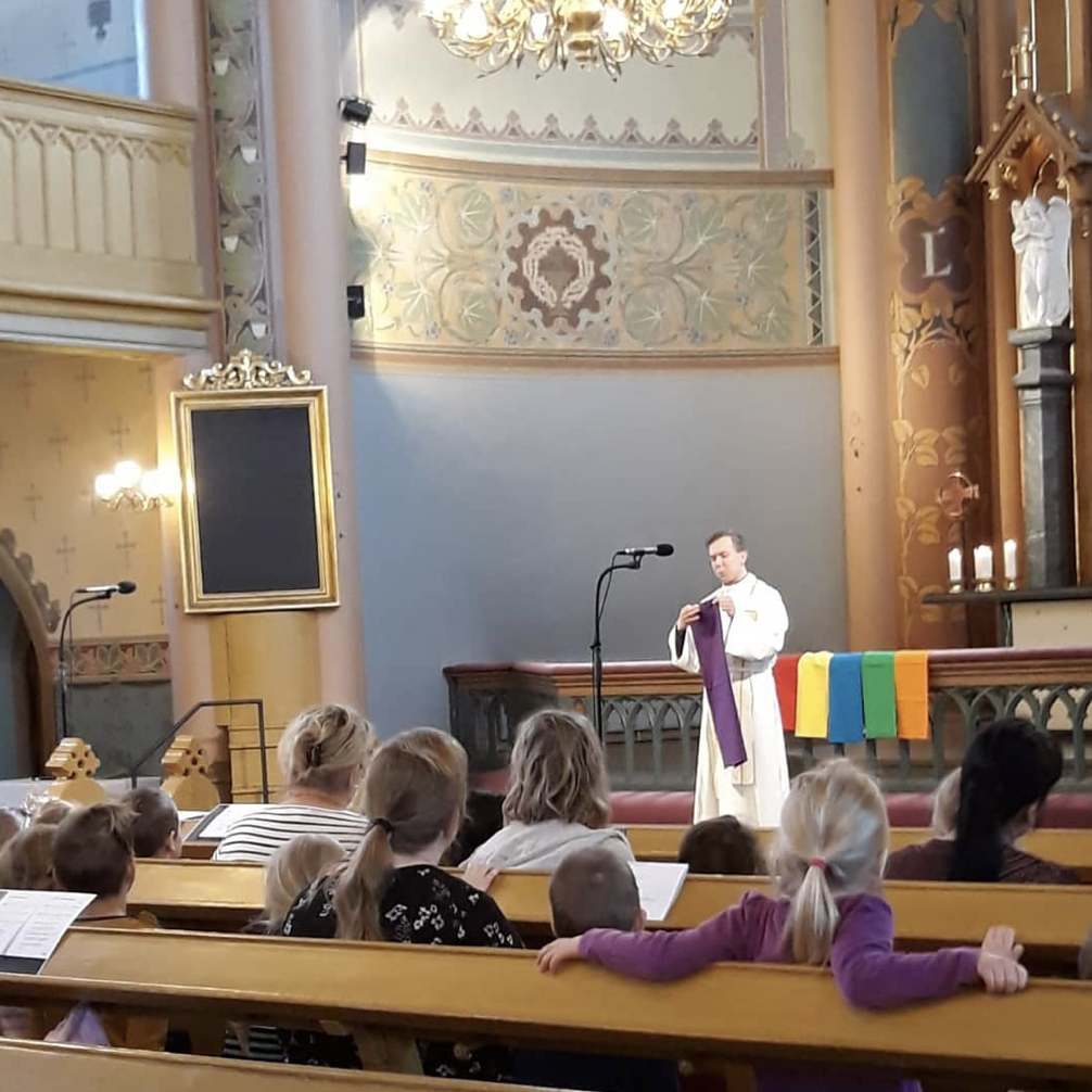 Kouluun lähtevien siunaaminen Kirkkohetki, jossa lapset seuraavat papin puhetta, jossa käsillä erivärisiä kankaita.
